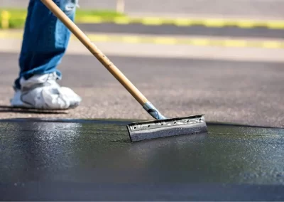 Person applying asphalt sealer on pavement with squeegee tool; wearing jeans and white sneakers.