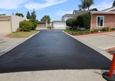 A freshly paved driveway stretches between two houses, its sleek surface catching the light. An orange traffic cone stands dutifully off to the side, hinting at recent work and raising unspoken asphalt sealcoating questions about durability and care.