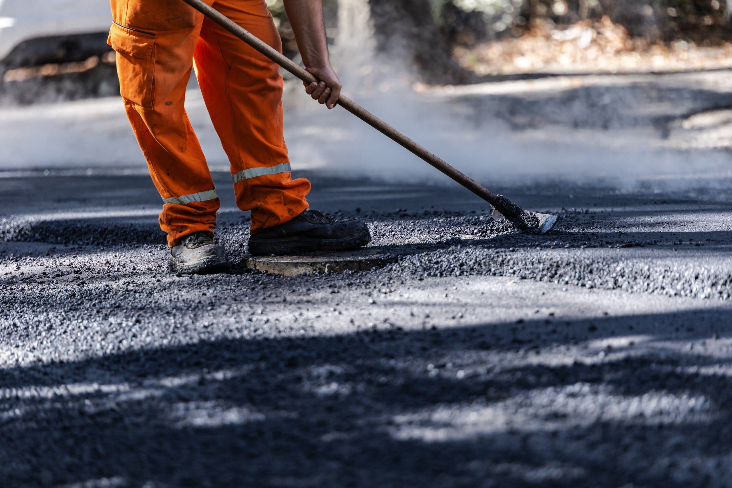 Worker repairing a road surface on a sunny day, wearing orange overalls and using a tool for asphalt application in a residential area Replacing vs. Resurfacing Asphalt Pavement
