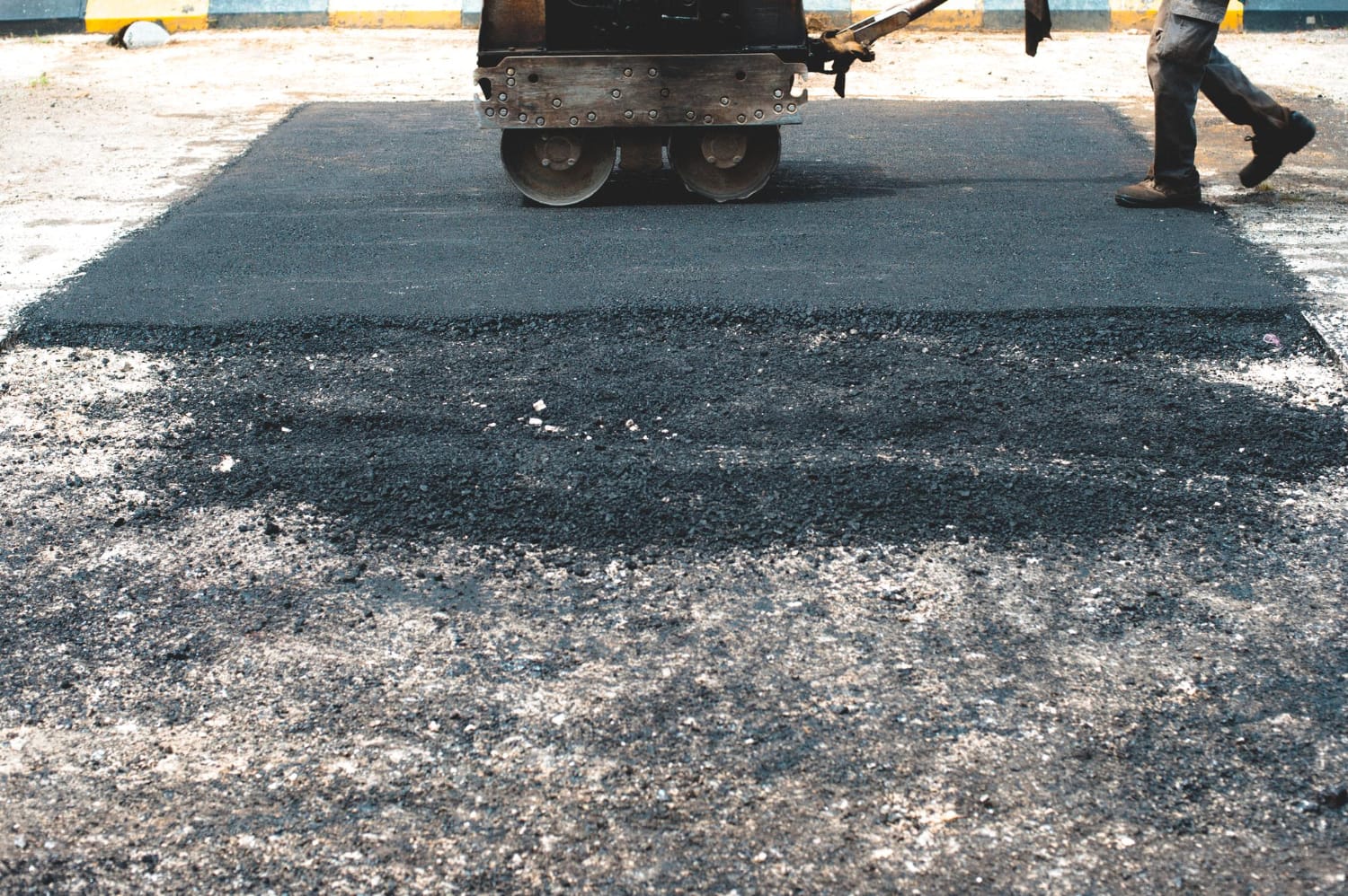 A road roller smooths fresh asphalt as a worker walks nearby on a construction site.